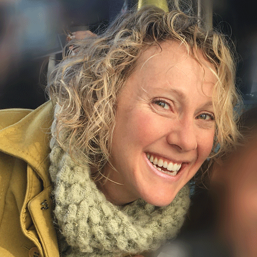 headshot of caucasian woman with short curly blond hair smiling. She is wearing an tan coat and a cream chunky scarf.